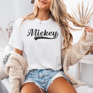 Puede incluir: Camiseta blanca con el nombre "Mickey" en negro, con una fuente desgastada. La camiseta tiene cuello redondo y mangas cortas. La persona lleva un pantal&oacute;n corto vaquero y un c&aacute;rdigan beige.
