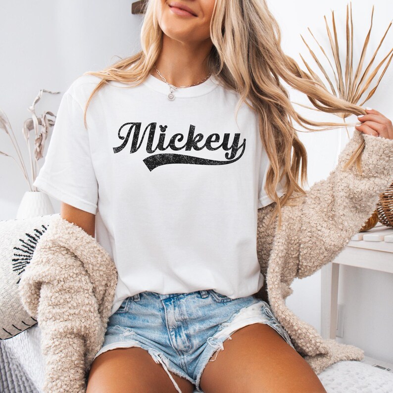 Puede incluir: Camiseta blanca con el nombre "Mickey" en negro, con una fuente desgastada. La camiseta tiene cuello redondo y mangas cortas. La persona lleva un pantal&oacute;n corto vaquero y un c&aacute;rdigan beige.