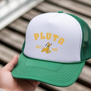 May include: A green and white trucker hat featuring the word "PLUTO" in yellow block letters. Below the word "PLUTO" is the text "EST 1930" and a cartoon image of Pluto. The hat has a green brim and mesh back.