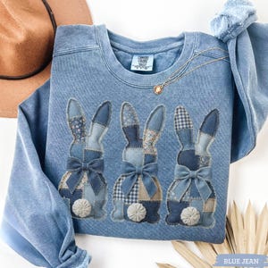 May include: Blue jean-colored sweatshirt featuring three appliqued bunny rabbits with patchwork patterns and bows. The bunnies have different patterns, including floral and gingham. A gold necklace and a brown hat are also visible.