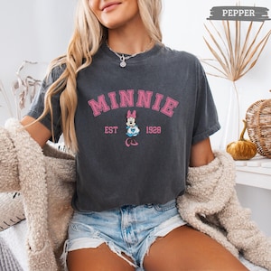 May include: Dark gray t-shirt with the word "MINNIE" in pink block letters. Below the word "MINNIE" is a graphic of Minnie Mouse and the text "EST 1928". The shirt is a vintage style.