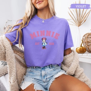May include: A violet t-shirt featuring the word "MINNIE" in pink block letters above a Minnie Mouse graphic. The graphic includes the text "EST 1928". The shirt is short-sleeved and made of a soft material.