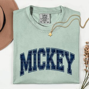 May include: A pale green t-shirt featuring the word "MICKEY" in a navy blue plaid pattern. A gold star pendant necklace and a brown hat are also visible. The shirt has a Comfort Colors label.