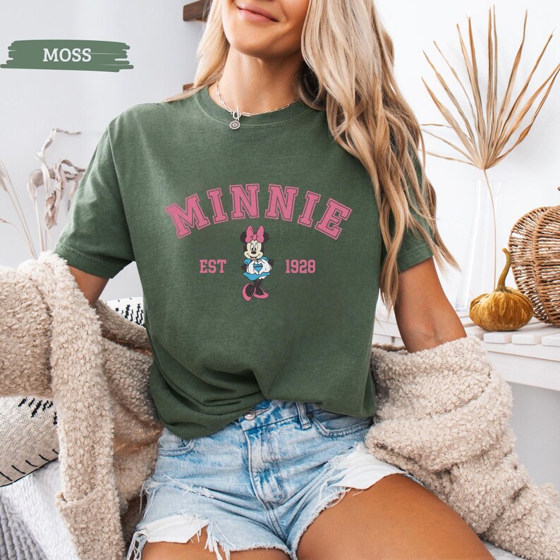 May include: Moss green t-shirt featuring the word "MINNIE" in pink block letters above a cartoon Minnie Mouse graphic. The text "EST 1928" is below the graphic. The shirt is a short-sleeved crew neck.
