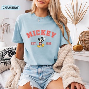 K&ouml;nnte beinhalten: Hellblaues T-Shirt mit dem Wort "MICKEY" in roten Blockbuchstaben &uuml;ber einem Cartoon-Bild von Mickey Mouse. Der Text "EST 1928" steht darunter. Das Wort "CHAMBRAY" befindet sich in der oberen linken Ecke.