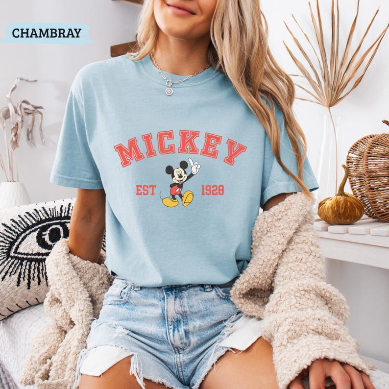 K&ouml;nnte beinhalten: Hellblaues T-Shirt mit dem Wort "MICKEY" in roten Blockbuchstaben &uuml;ber einem Cartoon-Bild von Mickey Mouse. Der Text "EST 1928" steht darunter. Das Wort "CHAMBRAY" befindet sich in der oberen linken Ecke.