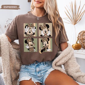 May include: Espresso-colored t-shirt featuring a four-panel design of Minnie Mouse in leopard print attire. Each panel showcases a different pose against a green and beige striped background. The shirt has a crew neck and short sleeves.