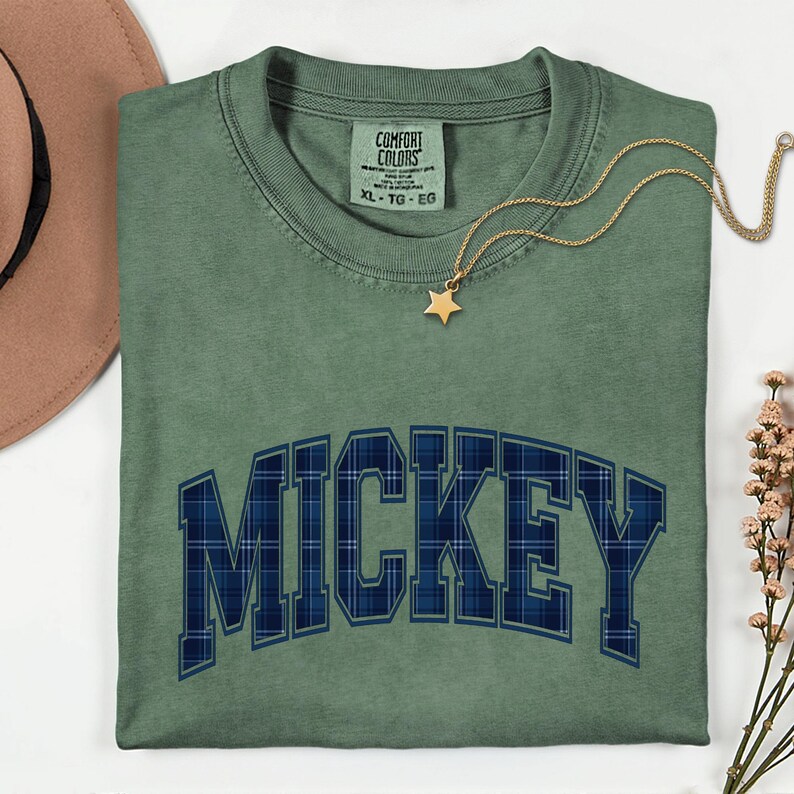 May include: Green t-shirt featuring the word "MICKEY" in a blue plaid pattern. A gold star pendant necklace and a brown hat are visible. The shirt has a Comfort Colors tag, size XL-TG-EG.