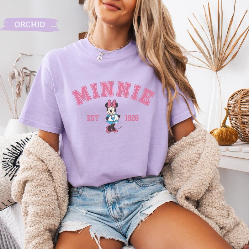 May include: A light purple t-shirt featuring the word "MINNIE" in pink arched lettering, with a Minnie Mouse graphic below. The text "EST 1928" is also printed on the shirt. The shirt is part of a clothing collection.