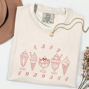 May include: Cream-colored t-shirt with the words "LAZY SUNDAES" in red, along with red illustrations of ice cream sundaes. A gold star pendant necklace and a brown hat are also visible.