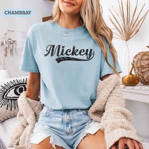 Puede incluir: Una camiseta azul claro con el nombre "Mickey" impreso en negro, con letras desgastadas. La camiseta de manga corta est&aacute; hecha de un material suave. La palabra "CHAMBRAY" est&aacute; impresa en un cuadro azul claro en la esquina superior izquierda.