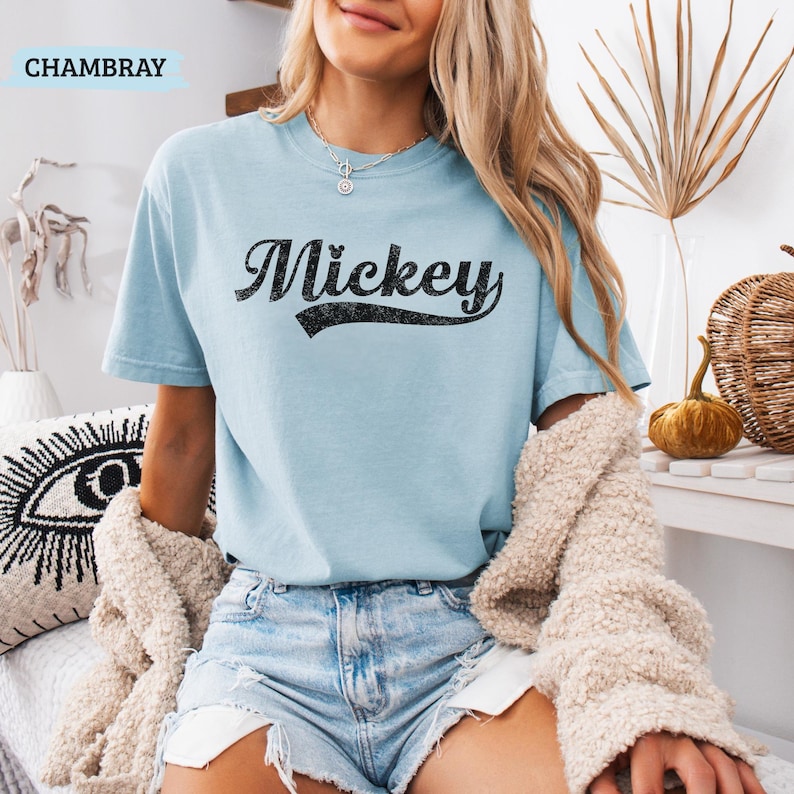 Puede incluir: Una camiseta azul claro con el nombre "Mickey" impreso en negro, con letras desgastadas. La camiseta de manga corta est&aacute; hecha de un material suave. La palabra "CHAMBRAY" est&aacute; impresa en un cuadro azul claro en la esquina superior izquierda.