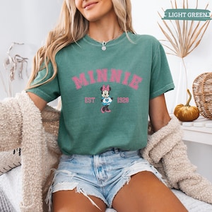May include: A light green t-shirt featuring the word "MINNIE" in pink, with "EST 1928" below and a Minnie Mouse graphic. The shirt is paired with denim shorts. The words "LIGHT GREEN" are in the top right corner.
