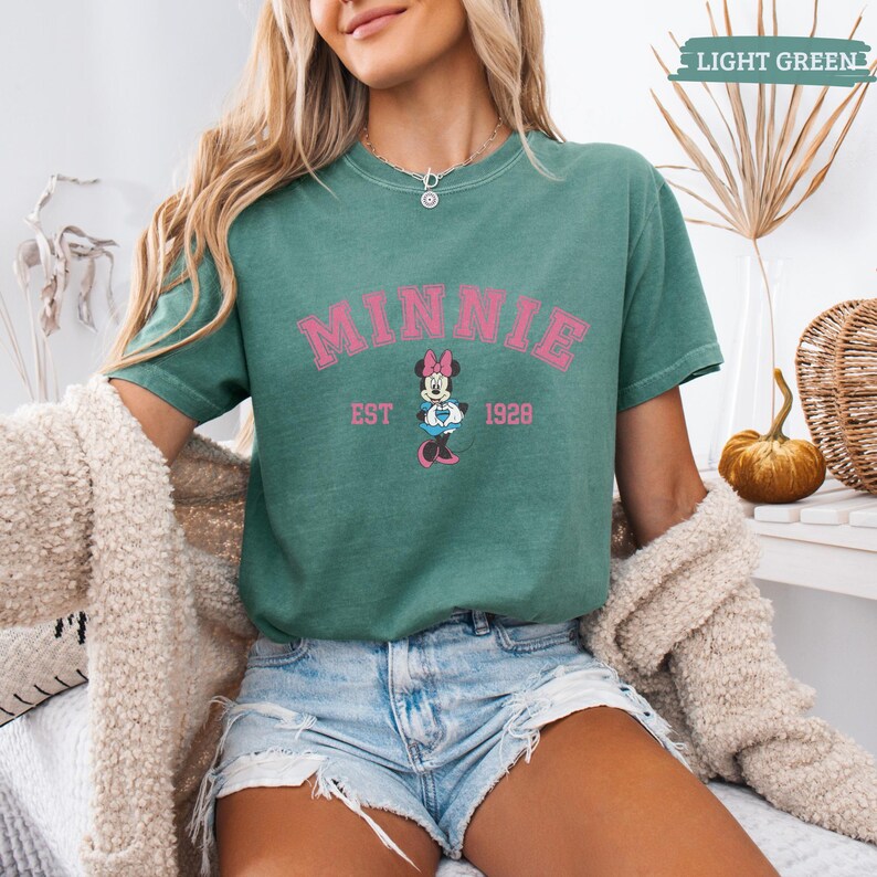 May include: A light green t-shirt featuring the word "MINNIE" in pink, with "EST 1928" below and a Minnie Mouse graphic. The shirt is paired with denim shorts. The words "LIGHT GREEN" are in the top right corner.
