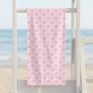 Puede incluir: Una toalla de playa rectangular con un patr&oacute;n de cuadros repetitivo en rosa claro y blanco. Cada cuadrado del patr&oacute;n contiene la silueta de una cabeza de rat&oacute;n con dos orejas redondeadas, orientada de forma consistente en el dise&ntilde;o.