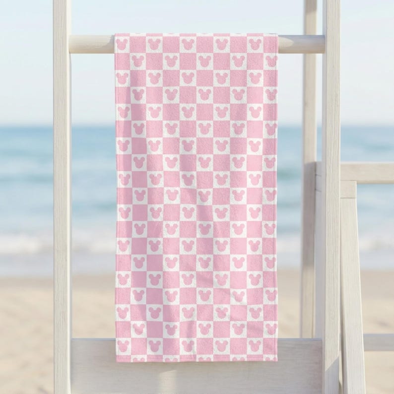 Puede incluir: Una toalla de playa rectangular con un patr&oacute;n de cuadros repetitivo en rosa claro y blanco. Cada cuadrado del patr&oacute;n contiene la silueta de una cabeza de rat&oacute;n con dos orejas redondeadas, orientada de forma consistente en el dise&ntilde;o.