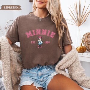 May include: Espresso-colored t-shirt featuring the word "MINNIE" in pink arched lettering, with a Minnie Mouse graphic below and "EST 1928". The shirt is short-sleeved and has a vintage, slightly faded appearance. The model is wearing denim shorts and a cream-colored cardigan.