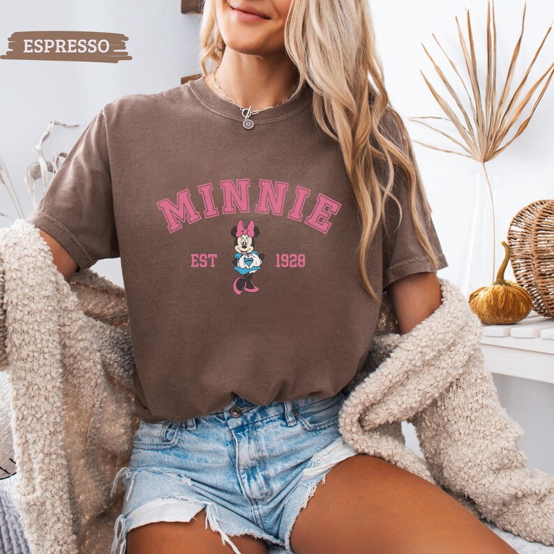 May include: Espresso-colored t-shirt featuring the word "MINNIE" in pink arched lettering, with a Minnie Mouse graphic below and "EST 1928". The shirt is short-sleeved and has a vintage, slightly faded appearance. The model is wearing denim shorts and a cream-colored cardigan.