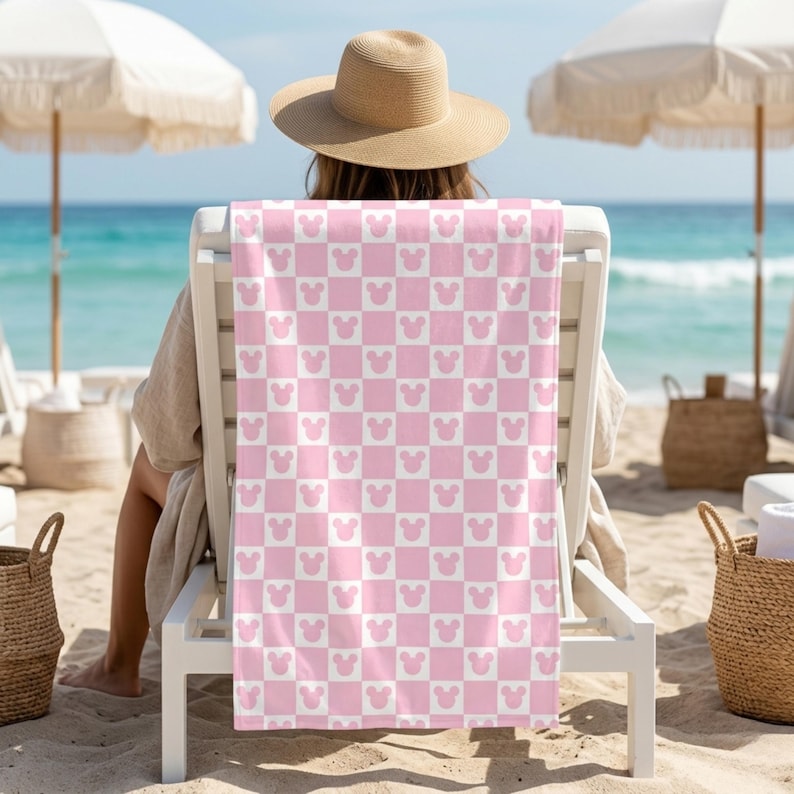 Puede incluir: Una toalla de playa rectangular con un patr&oacute;n de tablero de ajedrez repetitivo en rosa claro y blanco. Cada cuadrado rosa contiene una silueta blanca de un icono de cabeza de rat&oacute;n, mientras que cada cuadrado blanco contiene un icono de cabeza de rat&oacute;n rosa claro.