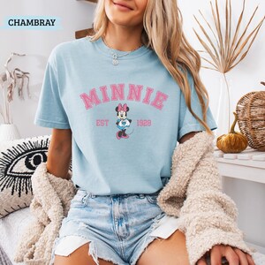 May include: Light blue t-shirt with the word "MINNIE" in pink block letters, and a Minnie Mouse graphic. The shirt also has the text "EST 1928". The word "CHAMBRAY" is in the top left corner.