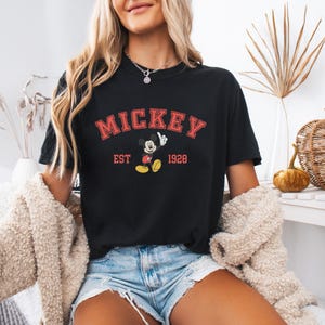 K&ouml;nnte beinhalten: Schwarzes T-Shirt mit dem Wort "MICKEY" in roter, bogenf&ouml;rmiger Schrift. Darunter befindet sich eine Grafik von Mickey Mouse und der Text "EST 1928". Das Shirt wird mit Jeansshorts und einer cremefarbenen Jacke getragen.