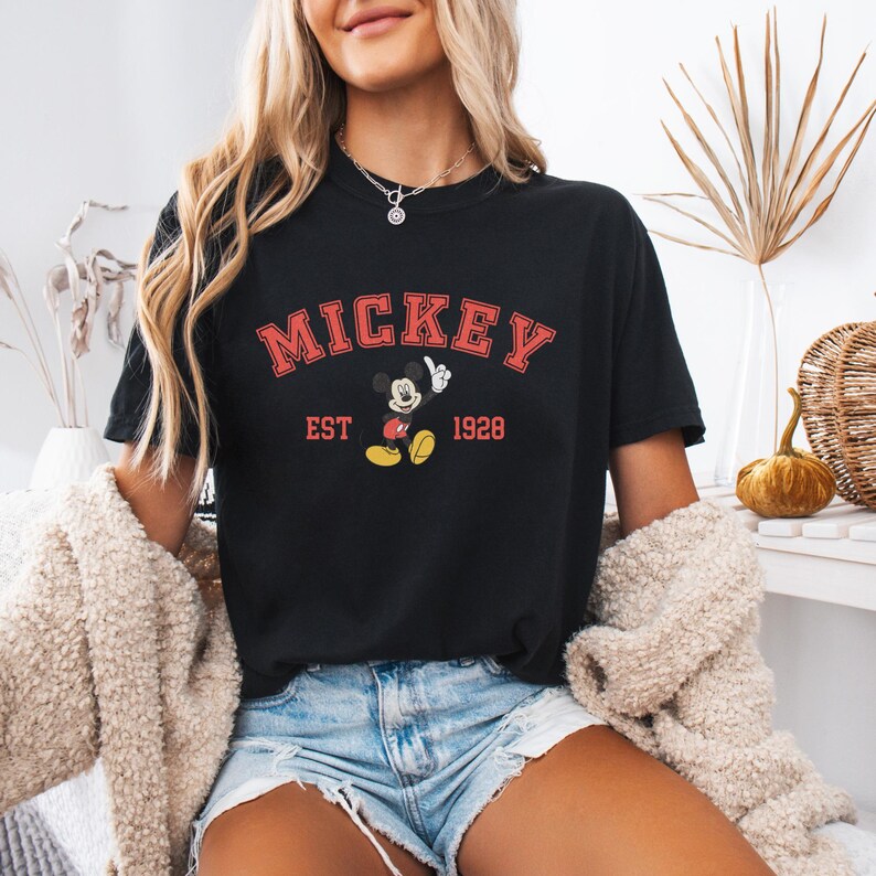 K&ouml;nnte beinhalten: Schwarzes T-Shirt mit dem Wort "MICKEY" in roter, bogenf&ouml;rmiger Schrift. Darunter befindet sich eine Grafik von Mickey Mouse und der Text "EST 1928". Das Shirt wird mit Jeansshorts und einer cremefarbenen Jacke getragen.