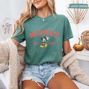 K&ouml;nnte beinhalten: Hellgr&uuml;nes T-Shirt mit dem Wort "MICKEY" in Rot, einer Mickey Mouse-Grafik und dem Text "EST 1928". Das Shirt ist ein l&auml;ssiges Oberteil mit kurzen &Auml;rmeln.