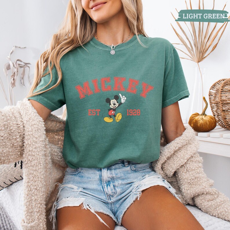 K&ouml;nnte beinhalten: Hellgr&uuml;nes T-Shirt mit dem Wort "MICKEY" in Rot, einer Mickey Mouse-Grafik und dem Text "EST 1928". Das Shirt ist ein l&auml;ssiges Oberteil mit kurzen &Auml;rmeln.