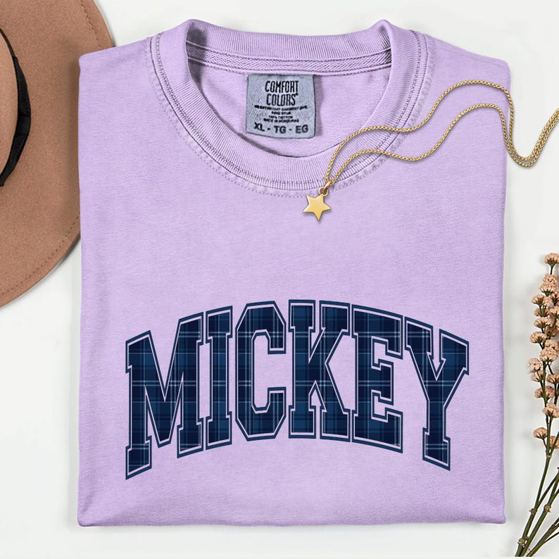 May include: A lilac t-shirt featuring the word "MICKEY" in navy blue plaid lettering. A gold star pendant necklace is draped across the shirt. The shirt is neatly folded, with a tag visible at the neckline.