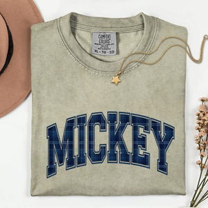 May include: A light green t-shirt featuring the word "MICKEY" in navy blue plaid lettering. A gold necklace with a star pendant is placed on the shirt. The shirt has a tag that reads "COMFORT COLORS" and the size "XL - TG - EG".