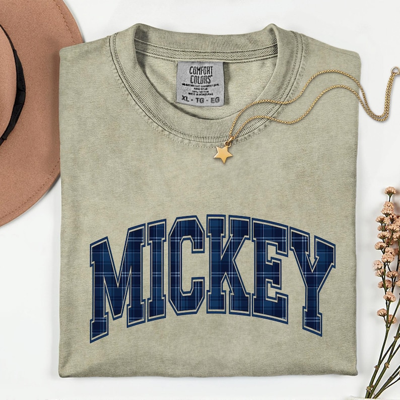 May include: A light green t-shirt featuring the word "MICKEY" in navy blue plaid lettering. A gold necklace with a star pendant is placed on the shirt. The shirt has a tag that reads "COMFORT COLORS" and the size "XL - TG - EG".