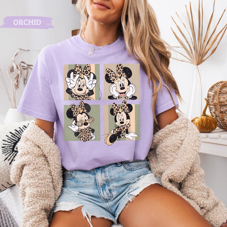 May include: A light purple t-shirt featuring a four-panel design of Minnie Mouse in various poses, each with a leopard print bow and outfit. The word "ORCHID" is printed at the top. The model is wearing denim shorts and a cream-colored cardigan.