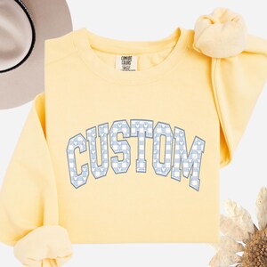 May include: A light yellow sweatshirt with the word "CUSTOM" in a blue and white gingham pattern. The letters have a Mickey Mouse head design. The sweatshirt is a Comfort Colors brand.