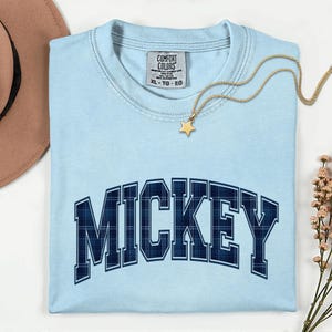May include: Pale blue Comfort Colors XL t-shirt featuring the word "MICKEY" in navy blue plaid lettering. A gold star pendant necklace and a sprig of pink flowers are also visible.