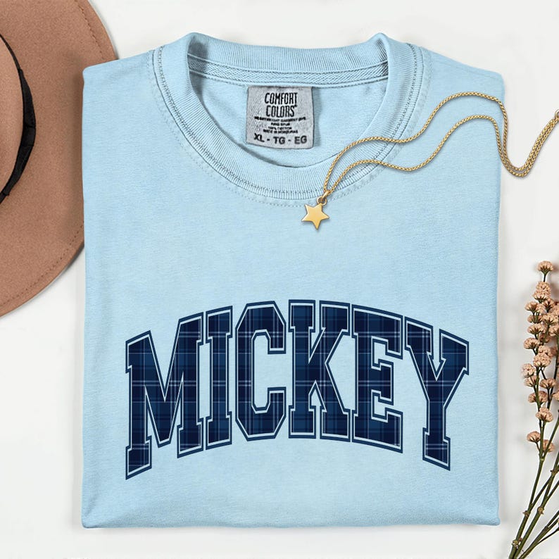 May include: Pale blue Comfort Colors XL t-shirt featuring the word "MICKEY" in navy blue plaid lettering. A gold star pendant necklace and a sprig of pink flowers are also visible.