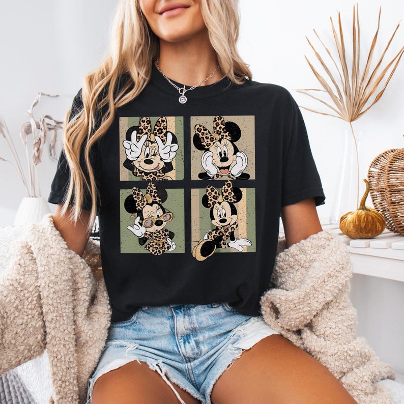 May include: Black t-shirt featuring a four-panel design of Minnie Mouse in various poses, each with a leopard print bow and accents. The background has a vintage, distressed look with green and tan stripes. A casual, comfortable tee.