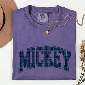 May include: A lilac Comfort Colors t-shirt featuring the name "MICKEY" in a blue plaid pattern. A gold star pendant necklace and a brown hat are also visible. The shirt is size XL-TG-EG.