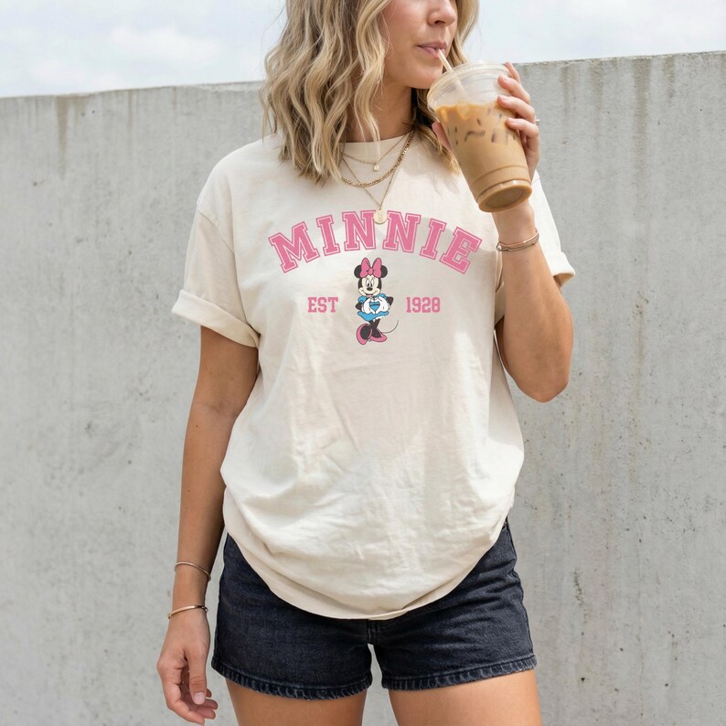 May include: Cream-colored t-shirt featuring the word "MINNIE" in pink, arched across the top. Below, a graphic of Minnie Mouse and the text "EST 1928" is printed. The shirt has rolled-up sleeves and is worn with black shorts.