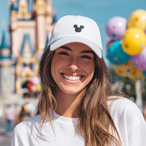 May include: A white baseball cap featuring a small, black, circular three-part icon embroidered on the front center. The cap has a structured crown, a curved brim, and a clean, minimalist design made from a solid white fabric material.