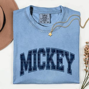 May include: A pale blue t-shirt featuring the name "MICKEY" in a navy blue plaid pattern. The shirt has a crew neck and short sleeves. A gold necklace with a star pendant is draped across the shirt. The shirt is a size XL-TG-EG.