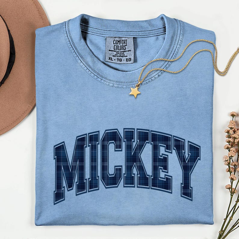 May include: A pale blue t-shirt featuring the name "MICKEY" in a navy blue plaid pattern. The shirt has a crew neck and short sleeves. A gold necklace with a star pendant is draped across the shirt. The shirt is a size XL-TG-EG.