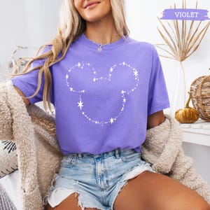 May include: A violet short-sleeved t-shirt featuring a white graphic outline of a mouse head silhouette composed of small dots and star shapes. The shirt has a crew neckline and a relaxed fit, with the word VIOLET printed in a small box at the top.