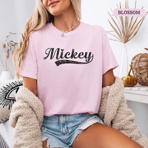 Puede incluir: Camiseta rosa claro con el nombre "Mickey" en escritura negra. La camiseta tiene mangas cortas y cuello redondo. La palabra "Blossom" est&aacute; impresa en una pancarta rosa en la esquina superior derecha.