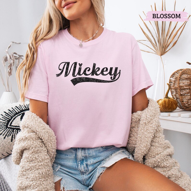 Puede incluir: Camiseta rosa claro con el nombre "Mickey" en escritura negra. La camiseta tiene mangas cortas y cuello redondo. La palabra "Blossom" est&aacute; impresa en una pancarta rosa en la esquina superior derecha.