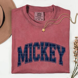 May include: A dusty rose Comfort Colors t-shirt with the word "MICKEY" in a blue plaid design. A gold star pendant necklace and a brown hat are also visible. The shirt is size XL-TG-EG.