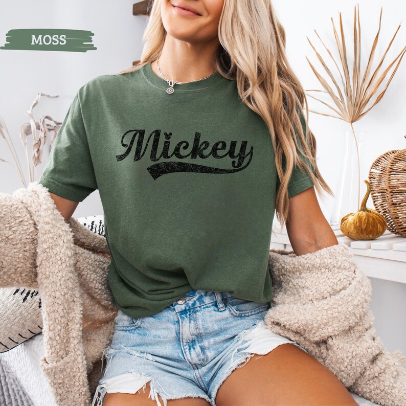 Puede incluir: Camiseta verde musgo con el nombre "Mickey" en escritura negra. La camiseta es de color liso y tiene mangas cortas. La palabra "MOSS" est&aacute; en un rect&aacute;ngulo verde en la esquina superior izquierda de la camiseta.