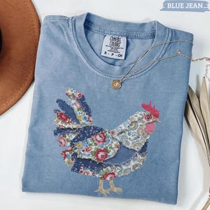 May include: A light blue Comfort Colors t-shirt featuring a patchwork chicken design with floral and paisley patterns. The shirt has a crew neck and short sleeves. A gold necklace with a pendant is draped across the shirt.