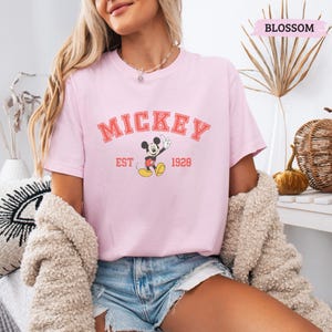 K&ouml;nnte beinhalten: Hellrosa T-Shirt mit dem Wort "MICKEY" in roten Blockbuchstaben &uuml;ber einem Cartoon-Bild von Mickey Mouse. Der Text "EST 1928" steht darunter. Das T-Shirt hat kurze &Auml;rmel.