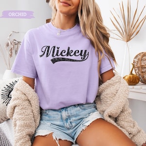 Puede incluir: Una camiseta de color lila claro con la palabra "Mickey" en escritura negra. La camiseta de manga corta tiene cuello redondo. La palabra "Orchid" est&aacute; en un rect&aacute;ngulo lila claro en la esquina superior izquierda.