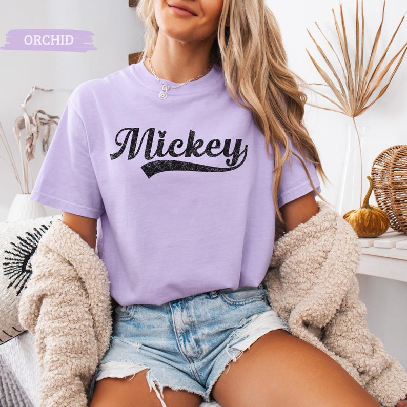 Puede incluir: Una camiseta de color lila claro con la palabra "Mickey" en escritura negra. La camiseta de manga corta tiene cuello redondo. La palabra "Orchid" est&aacute; en un rect&aacute;ngulo lila claro en la esquina superior izquierda.